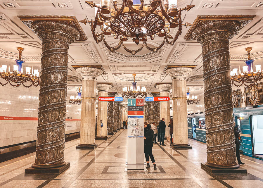 O femeie citeste o harta in interiorul statiei de metrou St Petersberg, cu candelabre atarnand de tavan si muluri de ipsos pe tavane si coloane.