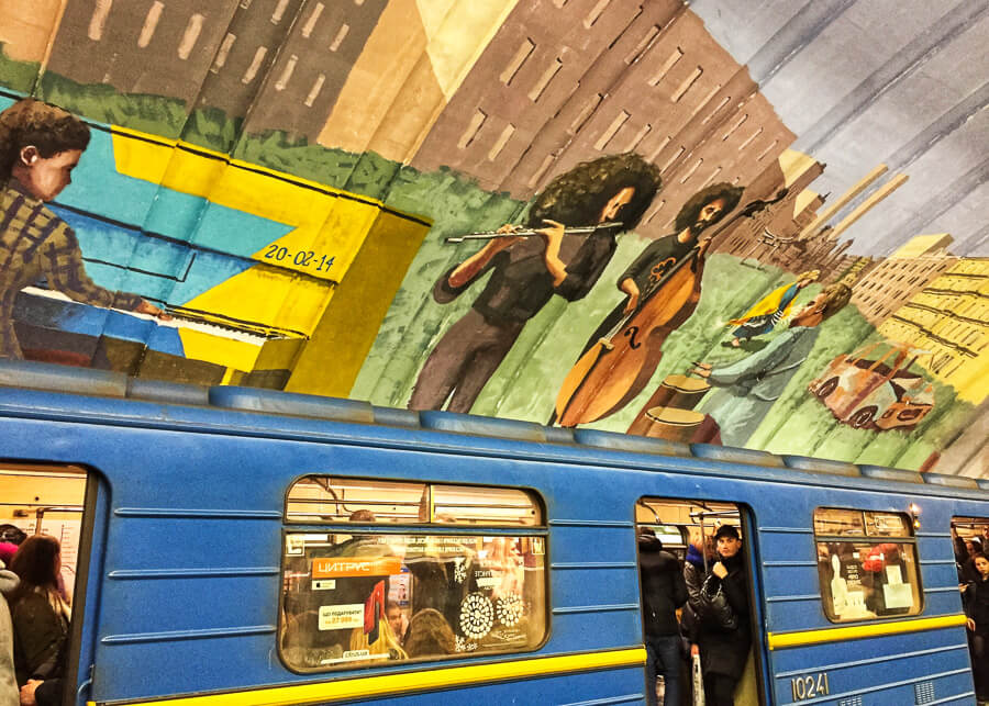 Un tren albastru trece printr-o statie de metrou din Kiev, cu picturi murale infatisand muzicieni pe acoperis.
