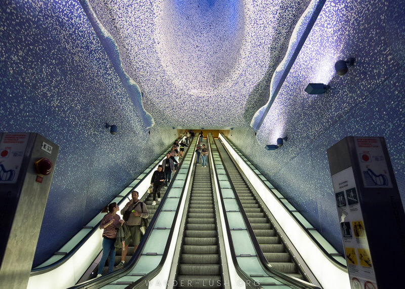 Scari rulante in interiorul unei statii de metrou subterane din Toledo din Napoli, cu placi albastre si albe pe pereti si tavan.